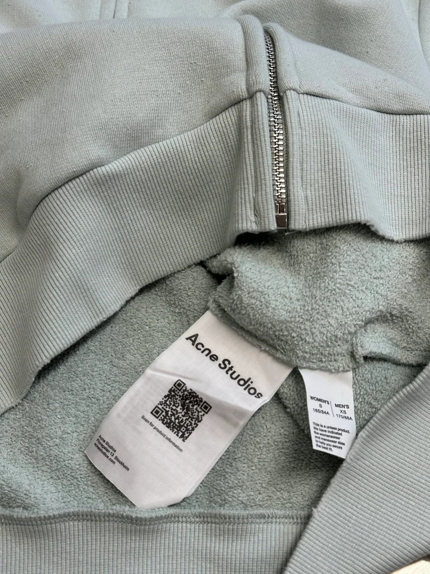 Ac vintage collared zipper sweatshirt uses 32-count top layer and 40-count bottom layer of 3-strand composite fleece structure, with a weight of g/㎡. The 32-count yarn on the top layer is fine and smooth, giving the surface a smooth touch and excellent anti-pilling properties; the 40-count 3-ply yarn on the bottom layer is treated with a raising process to form a rich and soft fluff layer, providing excellent warmth and skin-friendly wrapping. With 21 2×2 ribs, the g/㎡ weight ensures that the collar, cuffs and hem have excellent tensile fatigue resistance and shape retention capabilities. During piece dyeing, the entire fabric is immersed in the dye vat after weaving, and the dye molecules fully penetrate into the fibers, ensuring full, deep colors and excellent color fastness. Each batch of dyeing produces subtle differences, making the color tone of the single product unique. On this basis, the paint is sprayed irregularly on the surface of the garment through manual spray painting to create a splash-like retro effect. The distressed processing of holes causes the color to fade and mottle in parts. The ribbed trim is also treated to form slight whitening and fluffing, which visually echoes the spray-painted effect on the body, making the new clothes appear as if they have been worn for a long time and exposed to the sun. The front logo stamp uses a slurry printing process. The printing slurry contains a special microcapsule foaming agent. After being baked at high temperature, the slurry layer expands to form three-dimensional bulges, and the surface presents a granular texture similar to inkjet paint. This process simulates the effect of random drops of pigment after being atomized during spray gun spraying. The edges of the pattern present a natural feathering transition, rather than the rigid boundaries of glue printing. The printing surface has a fine graininess and soft elasticity to the touch, forming a closed loop of the process with the distressed effect of the spray painting on the body. size：xs s m l